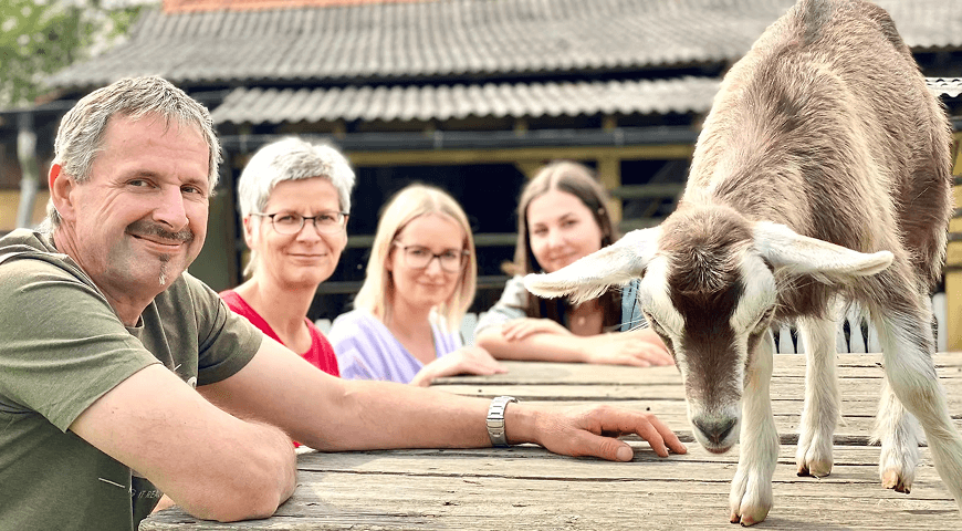 Eine Familie mit vier Personen steht neben einem Holztisch mit einer Ziege drauf