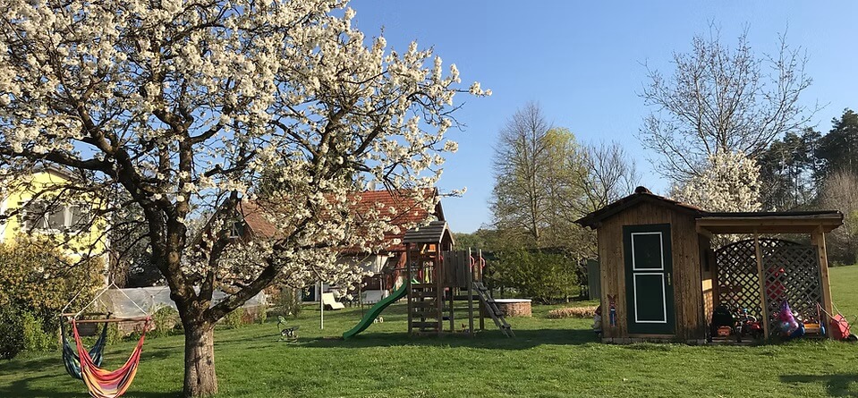 Grüne Wiese mit Spielplatz, Spielgeräten und Spielhaus. Hängesesseln hängen am blühenden Kirschbaum.