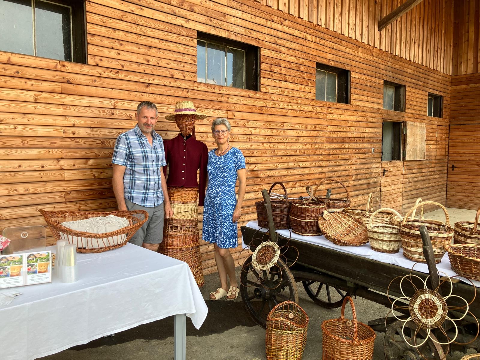 Ein Mann und eine Frau stehen neben einem Fuhrwagen mit Weidenkörbe. Dahinter ist ein schönes Gebäude mit Holzverschallung.v