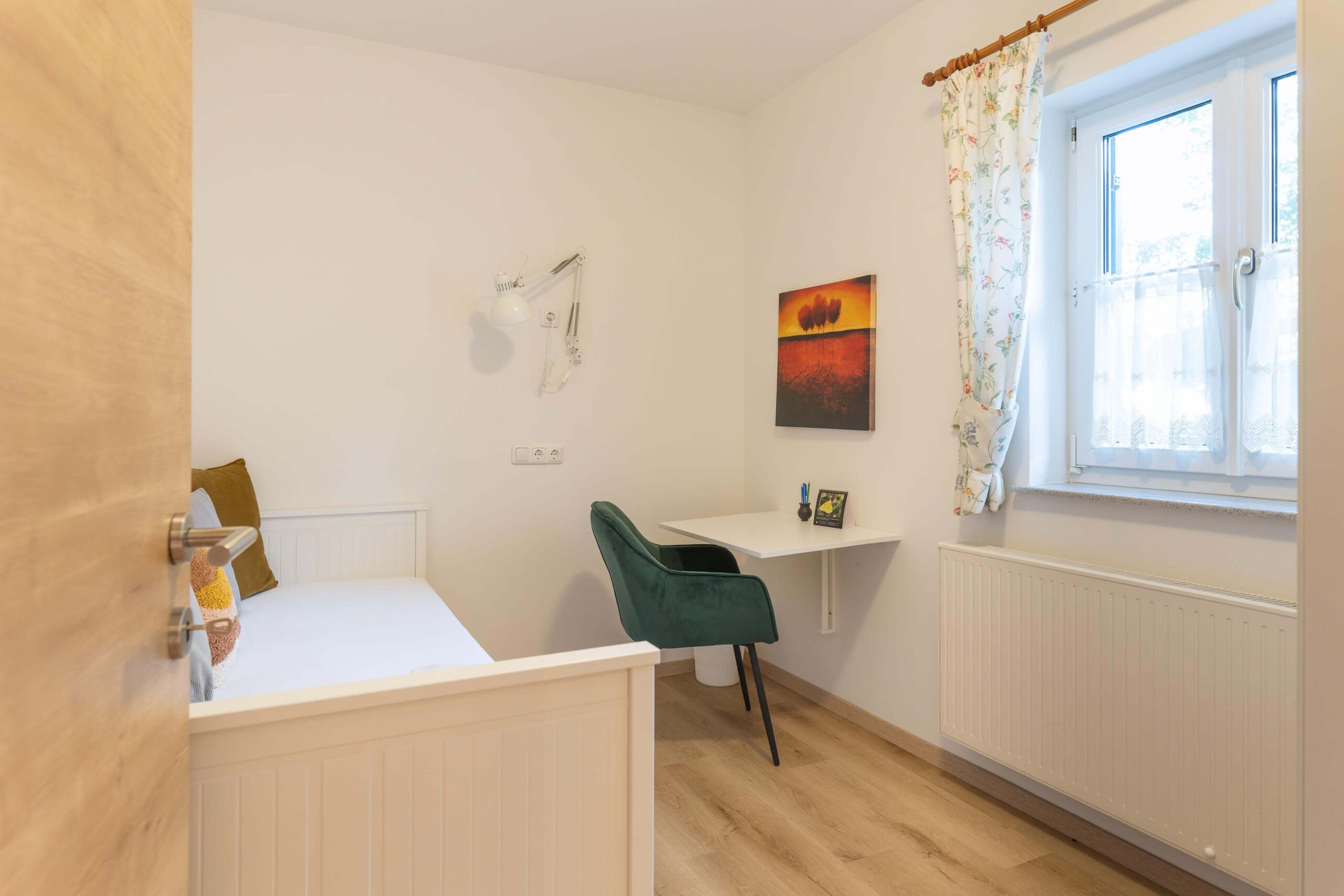 ein helles Zimmer mit Fenster, einem Bett, Schrank und Schreibtisch mit bequemen Stuhl.