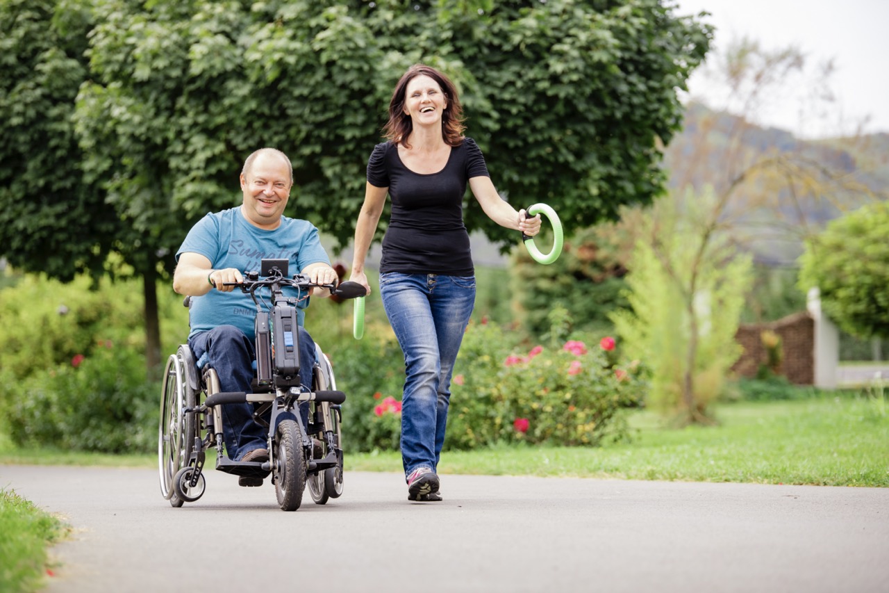 Ein Mann mit Handbike fährt den Weg im Landhaus Bender entlang eine Frau begleitet ihn. Beide wirken sehr Glücklich und entspannt
