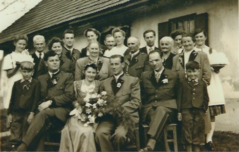 Schwarz-weißes Hochzeitsfoto mit Brautpaar in der Mitte, umgeben von einer großen Gruppe von Familienmitgliedern, die vor einem Gebäude posieren.