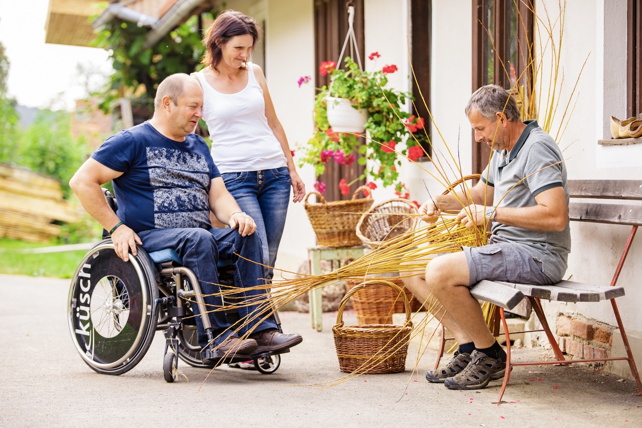 Ein Mann flechtet einen Weidenkorb und zwei Personen sehen dabei zu.