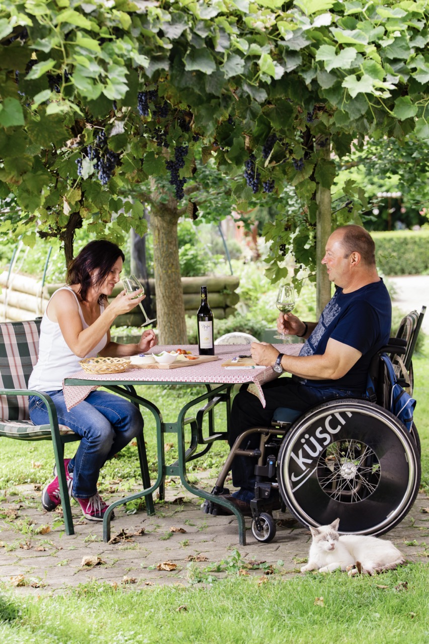 Eine Frau und ein Mann sitzen unter einer Weinlaube an einem schön gedeckten Tisch und trinken ein Glas Wein im Landhaus Bender.