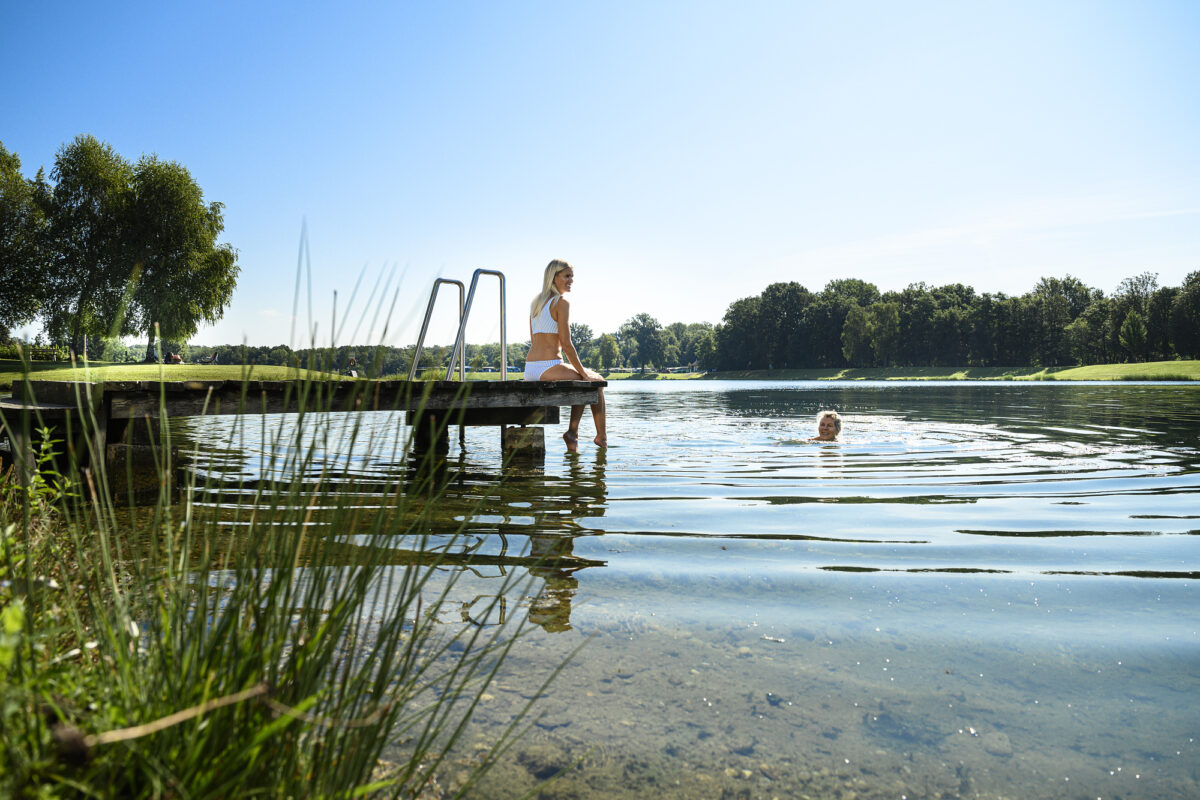 Frau sitzt auf einem Holzsteg am Badesee, während eine Person im klaren Wasser schwimmt – Sommerurlaub in der Thermen- und Vulkanland-Region.