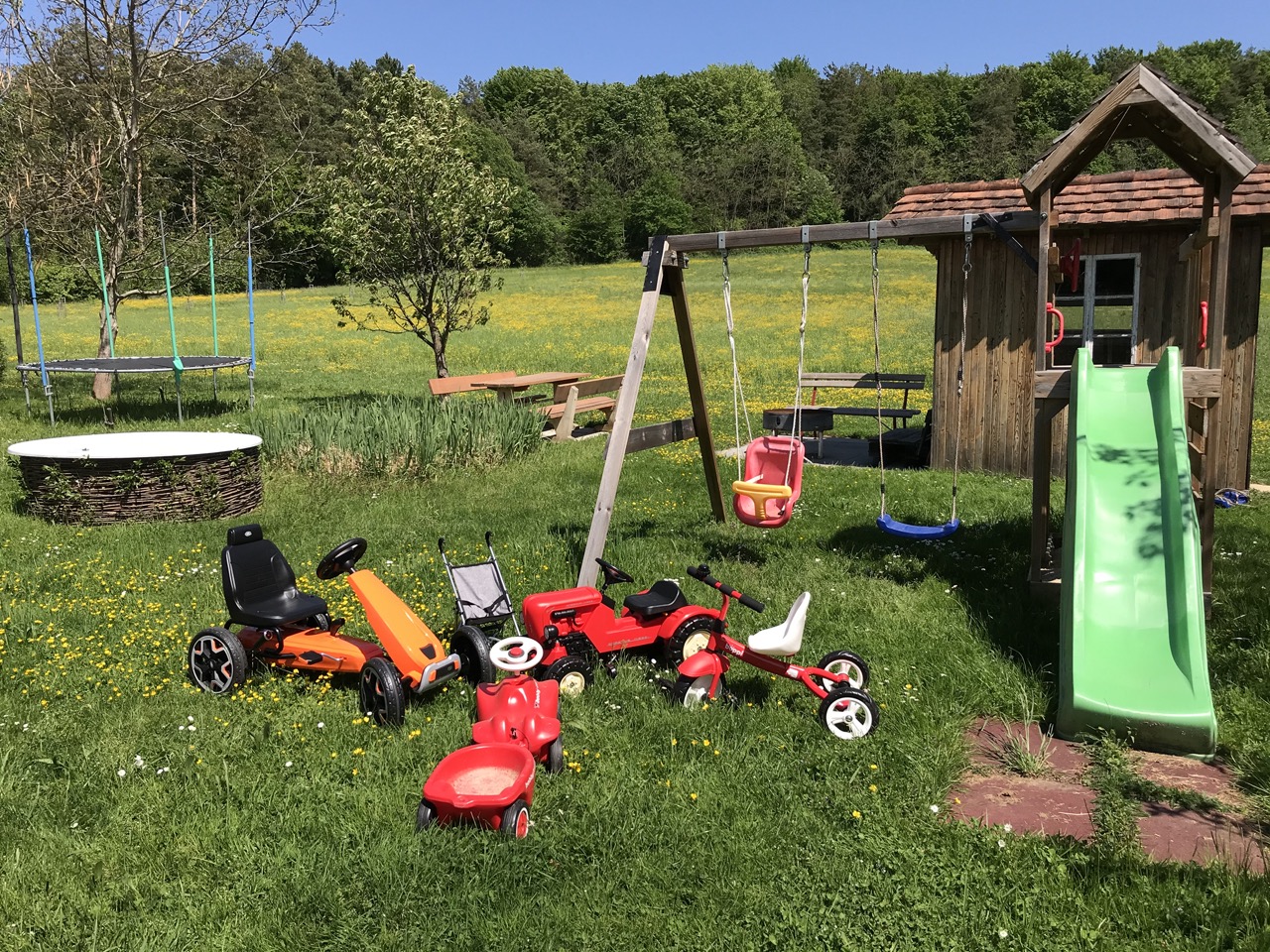 Großer Spielplatz mit Rutsche, vielen Spielgeräten und Fahrzeuge in unberührter Naturwiese. Dahinter ein schöner Laubwald.