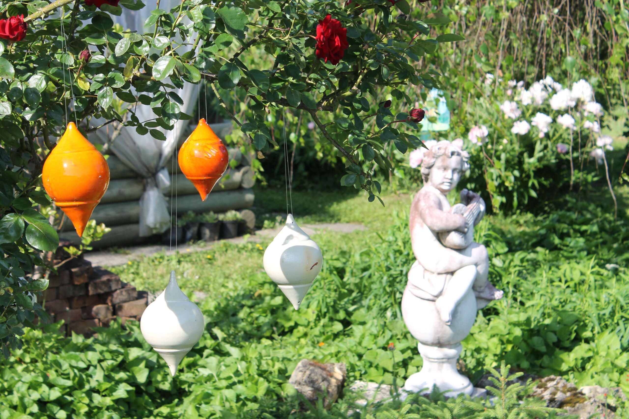 Geschmackvolle Gartengestaltung mit hängender bunter Keramik. Daneben eine weiße Figur aus Keramik.