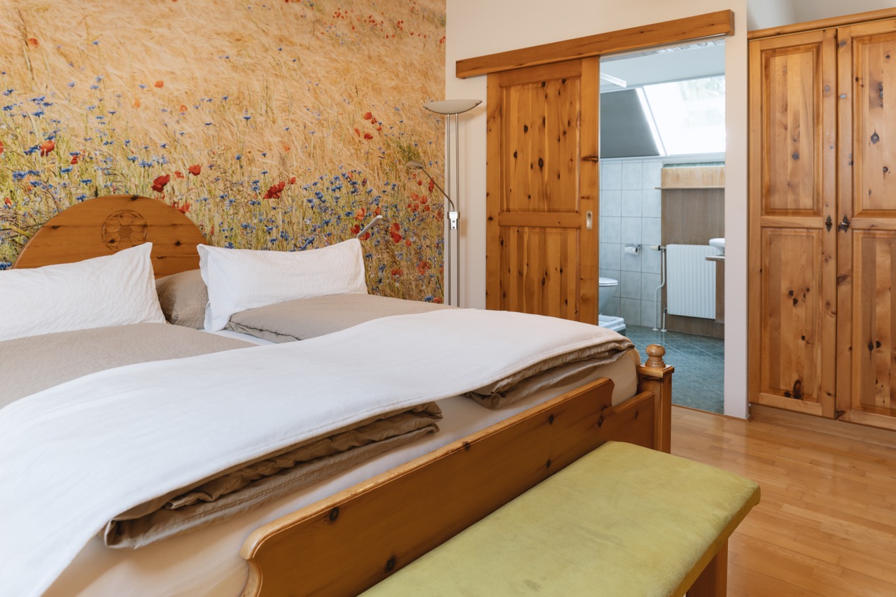 Doppelbett in Zirbenholz, Wandtapete mit Kornblumen und Mohn. Direkter Zugang in das schöne Badezimmer mit Fenster