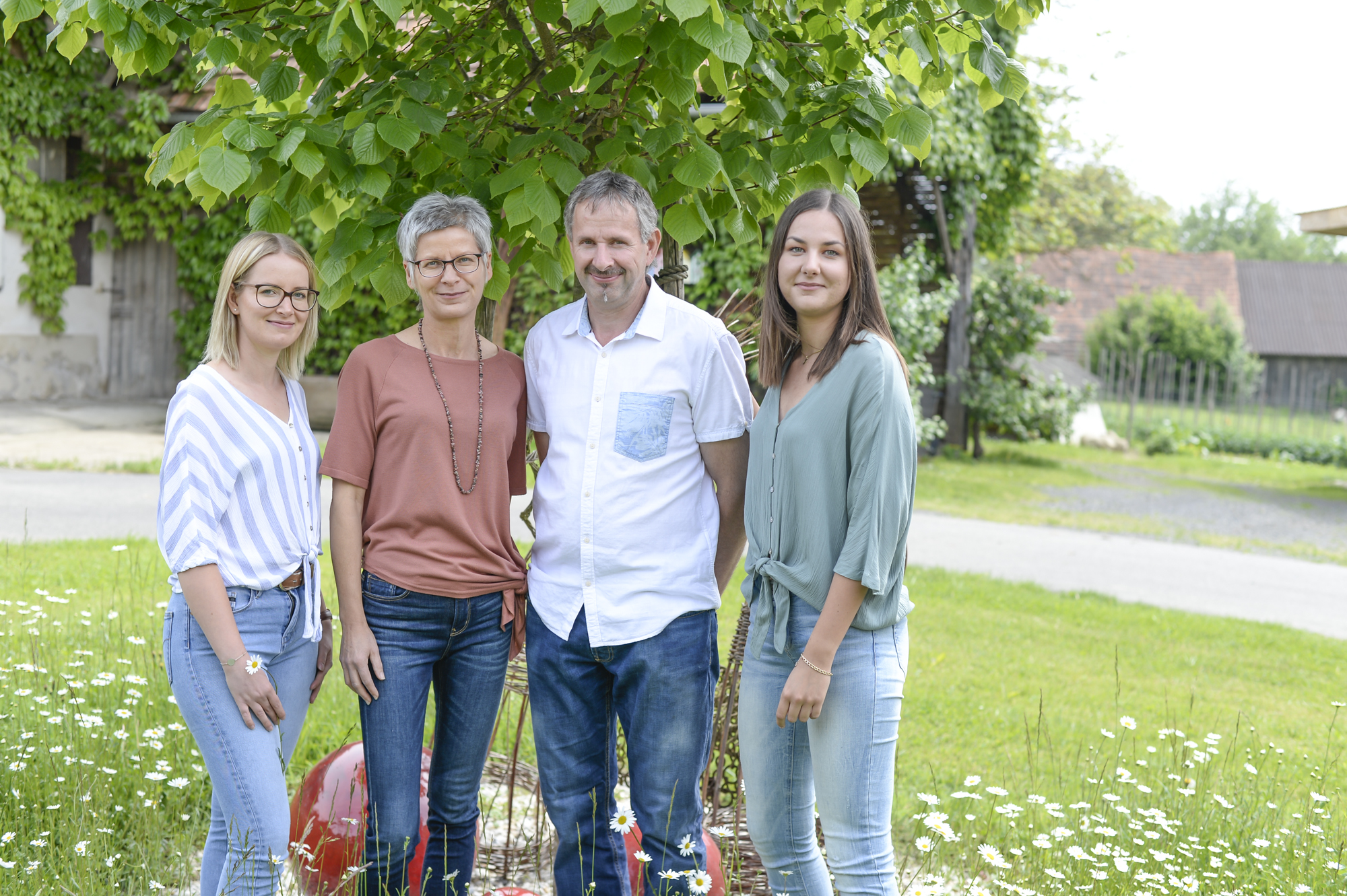 Eine Familie mit vier Personen steht in Innenhof eines Bauernhofes vor einem Lindenbaum und lächelt in die Kamera